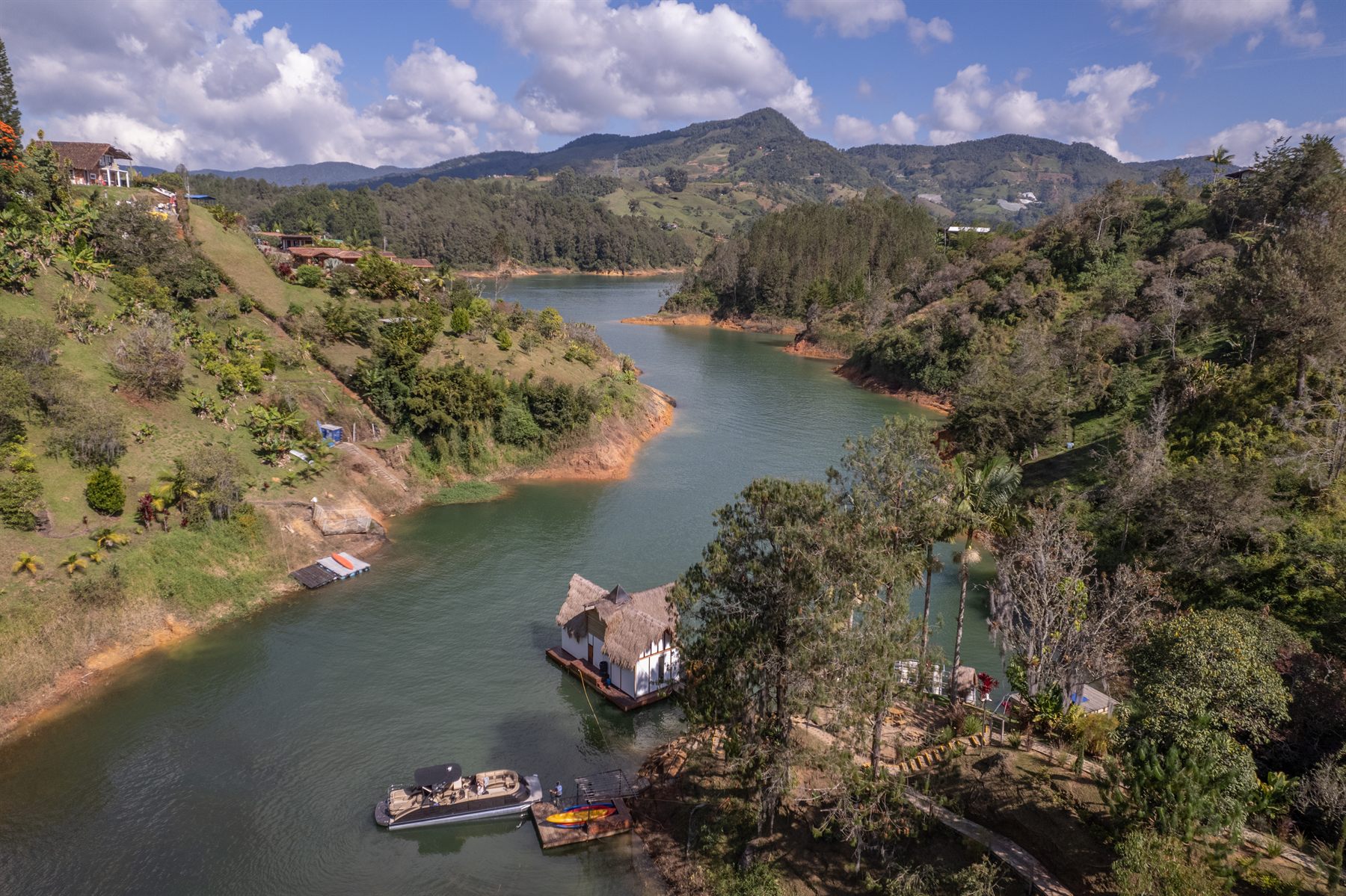 Vista aérea con drone del embalse de Guatapé junto a IBUKU, con muelle flotante de techo de paja y lancha, rodeado de montañas verdes
